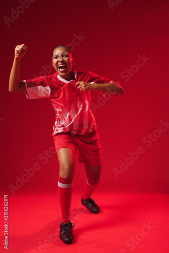 Female athlete shouting, raising fist, pulling jersey in studio with red jersey, black cleats
