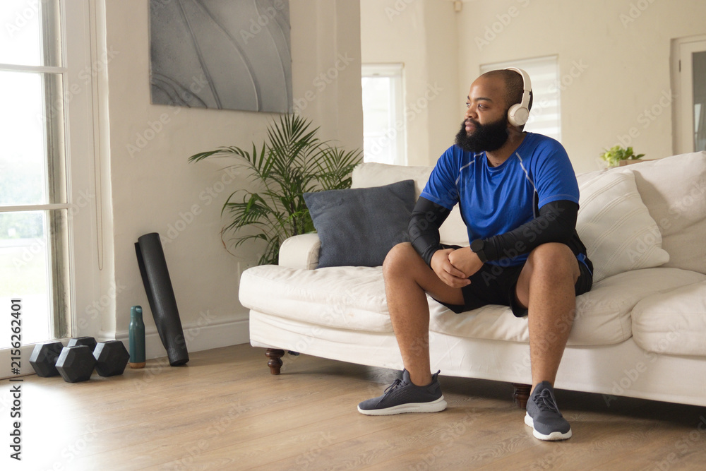 Fototapeta premium African American adult man sitting on sofa wearing headphones and blue shirt, dumbbells, copy space