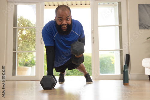 African American man performing renegade row in blue shirt by glass doors with dumbbell and bottle