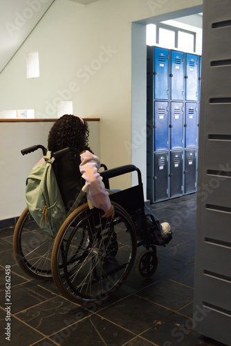 Manual wheelchair is sitting in school corridor near blue lockers, mint backpack hanging