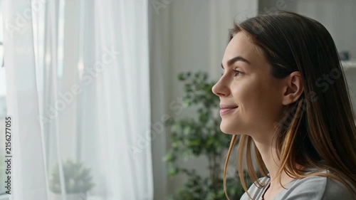 Side view profile of happy young woman smiling while looking out a bright window with white curtains in a sunlit interior.