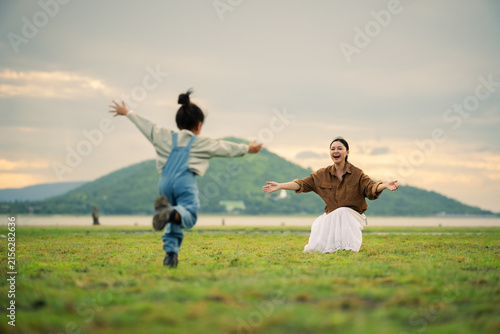 excited preschool child girl running into her mother's open arms on grass field near the river with mountain
