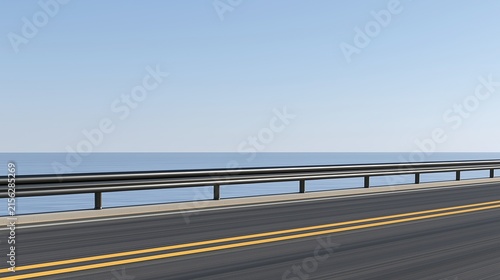 Empty coastal asphalt road with double yellow lines and black metal guardrail next to calm blue sea under clear light blue sky