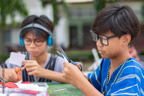 Two Asian teenage boys concentrating on cutting paper with scissors, doing arts and crafts or a school project together outdoors.