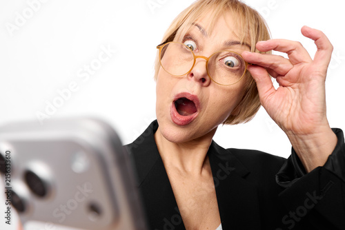 Obraz na płótnie Surprised blonde woman in business suit wearing glasses reacts to smartphone on white background, open mouth and raised fingers, shocked expression, close up dramatic portrait face