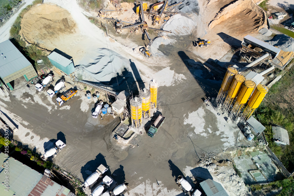 Fototapeta premium A busy concrete plant thrives with trucks and machinery moving around. Yellow silos tower over piles of gravel and sand under the bright midday sun