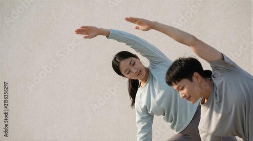 A young Asian couple practicing yoga and stretching exercises together outdoors against a minimalist neutral wall.