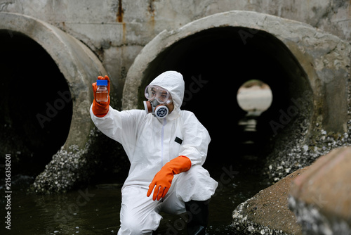 Scientist in hazmat suit tests polluted water near concrete drainage pipes, holding a device while kneeling in contaminated runoff to monitor wastewater and environmental hazards.