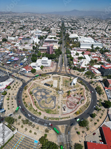 Aerial drone view captures the vast urban landscape of Guadalajara, Mexico, featuring the iconic Normal Roundabout and bustling city streets under a clear sky.