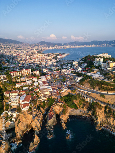 Stunning drone aerial view showcasing Acapulco's famous La Quebrada cliffs, coastal city, and vast bay during beautiful golden hour. The vibrant urban landscape meets the rugged Pacific coastline