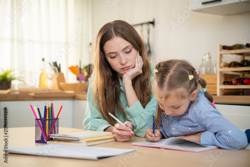 Tired mother sitting at kitchen table looking stressed while her young daughter doing homework with color pencils. Exhausted mother or tutor looking bored while helping little girl with home schooling