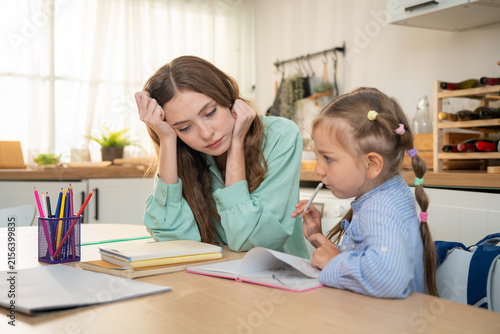 Tired mother sitting at kitchen table looking stressed while her young daughter doing homework with color pencils. Exhausted mother or tutor looking bored while helping little girl with home schooling