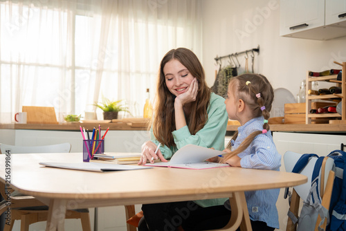Happy mother smiling while looking at her cute daughter reading notebook at home table. Young mother enjoying home learning time and supporting her happy daughter with school homework in kitchen.