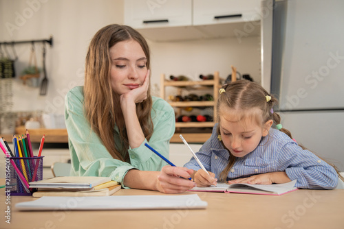 Mother resting chin on hand while watching her focused daughter write homework at home. Young mother sitting next to little girl doing homeschooling exercise with colored pencils at kitchen table.