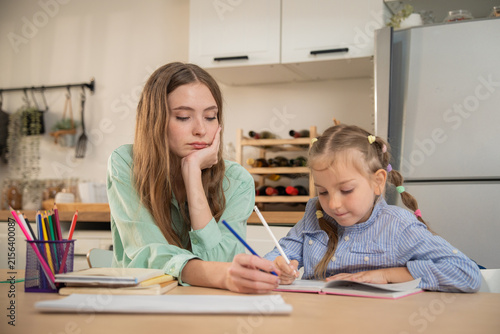 Mother guiding her little daughter's hand while writing homework at home kitchen table. Young mother helping daughter write in notebook during home study, showing care and patience despite being tired