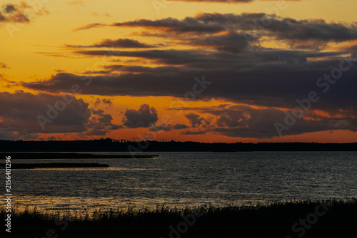 Konstfotografi Sunset over calm water with silhouetted shoreline and clouds reflecting warm hue