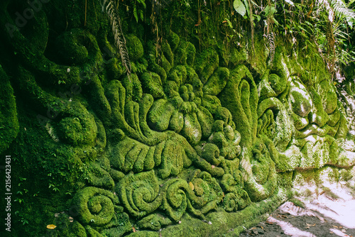 Ancient stone carving relief covered in vibrant green moss, revealing intricate Balinese art details and cultural heritage, thriving within a tropical jungle setting