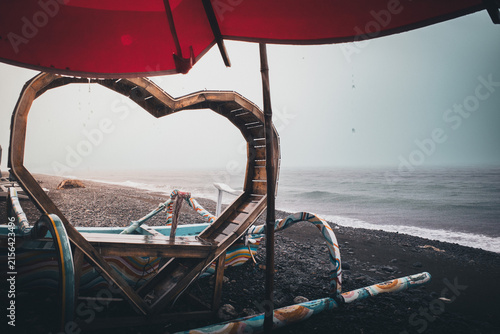 Heart-shaped boat structure on a black sand beach, with rain falling on the ocean under a grey sky, creating a melancholic and romantic atmosphere of solitude