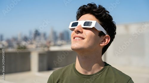 Young man wearing solar eclipse glasses smiling on city rooftop