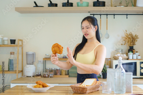 Canvas Print Fit Asian woman in yellow sports bra gestures refusal toward fried chicken in a modern kitchen, representing dieting, willpower, and healthy lifestyle choices