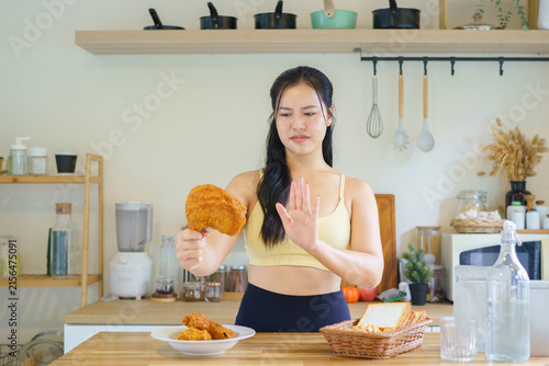 Obraz na plátně Fit Asian woman in yellow sports bra gestures refusal toward fried chicken in a modern kitchen, representing dieting, willpower, and healthy lifestyle choices