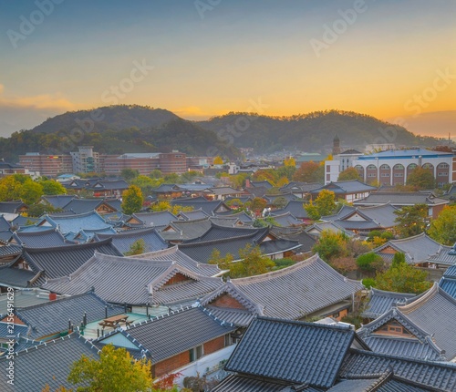 a panoramic view of a traditional Korean village with distinctive tiled roofs under a warm, hazy sky at sunset. The mountains in the background add depth to the scene. in Jeonju, South Korea