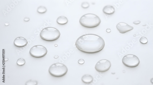 Clear water droplets of various sizes scattered on a smooth, light surface, reflecting ambient light.