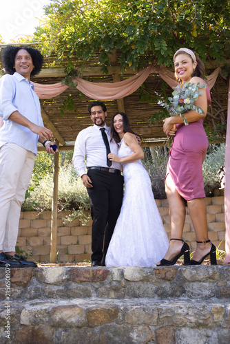 Bride in white gown, groom in black tie, bridesmaid holding bouquet posing on steps under pergola
