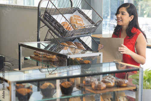 Mid-adult woman wearing red top packing pastries into brown paper bag at bakery counter, copy space