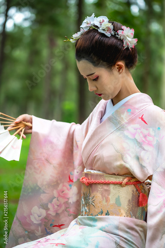 Fotografie Graceful young Japanese geisha in pastel kimono enjoying a vibrant green garden while holding folding fan