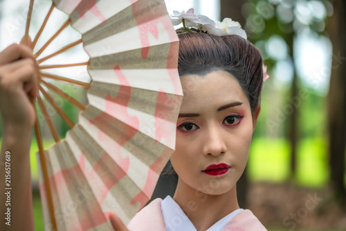 Portrait young Japanese geisha in a delicate pastel kimono holding a traditional fan covering face enjoying vibrant green garden 