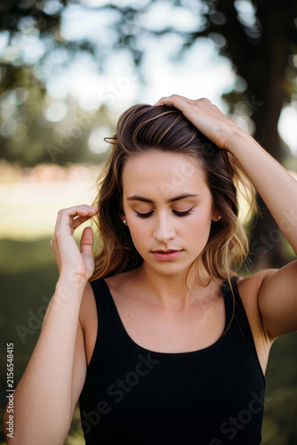 A serene young woman with long brown hair standing in a lush park, touching her hair with eyes closed in a moment of peaceful reflection under bright, natural sunlight.