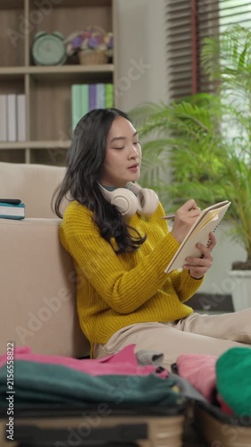 Asian woman writing checklist before packing suitcase for vacation trip