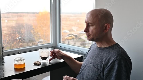 Middle aged man drinking coffee and eating cookies with honey while relaxing by a window with an autumn view in his apartment