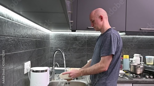 Bald man washing dishes with a sponge and running water in a modern dark grey kitchen sink after dinner