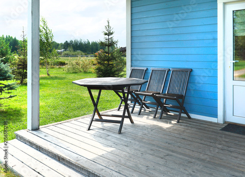 Wooden veranda with dark folding table and chairs by blue cabin. Cozy slow living ecotourism outdoor lifestyle concept. Peaceful summer park garden family vacation nature background.
