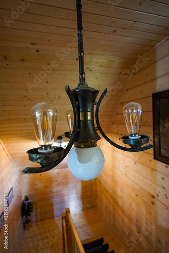 Vintage metal chandelier with exposed filament bulbs hangs in a warm wood-paneled interior above a staircase, creating a cozy rustic atmosphere for hospitality and interior design themes.