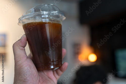 Hand holding iced black coffee or Americano coffee in plastic cup, against in coffee shop background