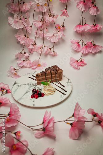 Layered chocolate cake slice with cream filling and berries on a square white plate framed by blooming sakura branches