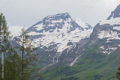 Picturesque Grossglockner High Alpine Road in Austria surrounded by rugged snowy peaks, green Alpine slopes and dramatic cloudy mountain scenery.