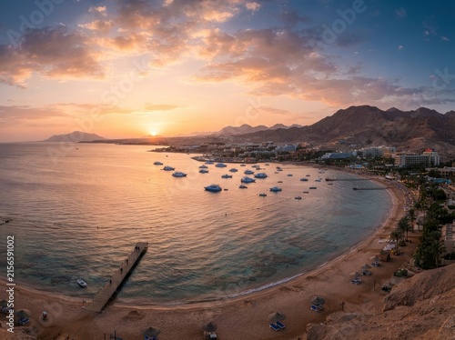 A panoramic high-angle view of a tropical coastal resort at sunset, featuring a sandy beach, a long wooden pier, anchored boats in the calm sea, and mountain ranges under a colorful cloudy sky.