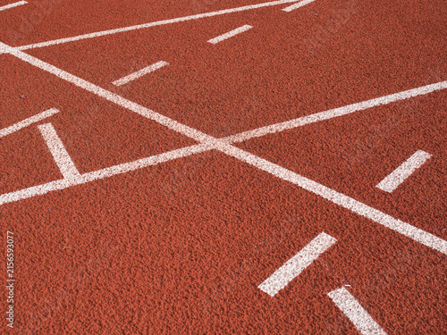 Photography Textured red rubber running track surface featuring solid white lane lines contrasted with smaller blue and yellow markings