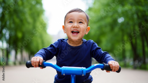 A happy young Asian boy laughing while riding his blue bicycle in a lush green park, capturing the pure joy of childhood activity, outdoor exercise, and early learning during a bright day.