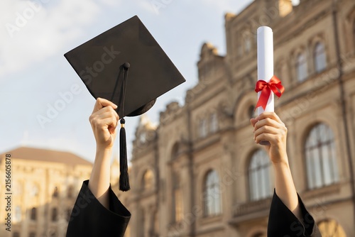 Close up of graduate hands raising cap and diploma against building backdrop