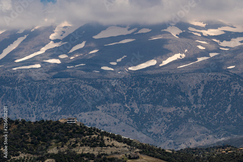 Bild auf Leinwand A lonely building sits on a green hillside before a majestic snow-capped mountain range under heavy clouds, creating a breathtaking scenic landscape view