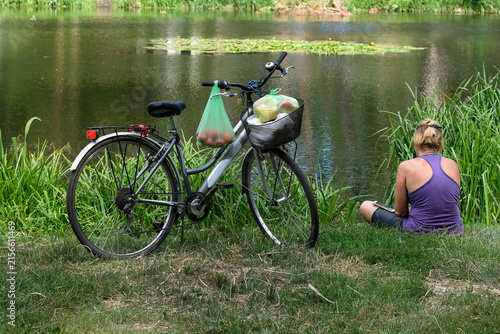 Young girl sits by a pond in sunny summer. Nearby is a bicycle with products. Outdoor recreation.