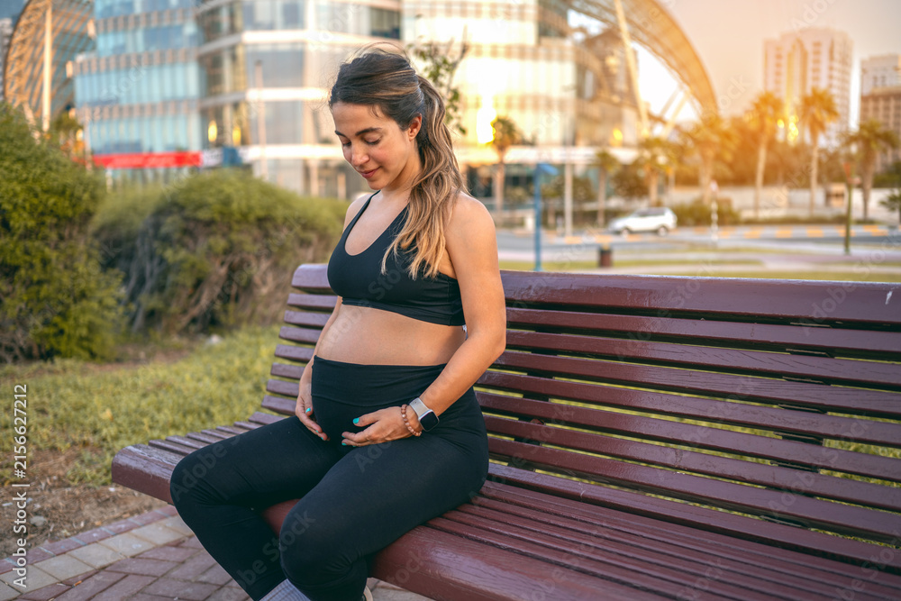 Fototapeta premium Pregnant woman in sportswear sitting on a park bench and looking down at her belly