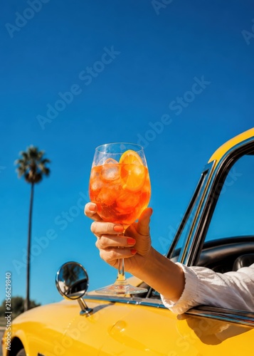 Refreshing orange spritz cocktail held from yellow vintage convertible on sunny summer day