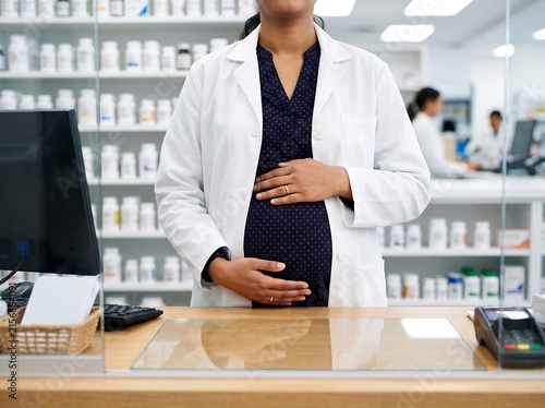 Pregnant pharmacist in white lab coat standing at pharmacy counter, expectant healthcare professional working in drugstore, medical worker holding baby bump in clinic setting