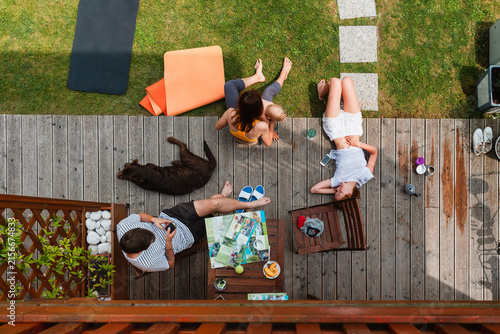 A young family relaxes on a wooden patio deck and green lawn in their backyard while a dog sleeps nearby. Authentic summer weekend lifestyle concept showing togetherness and outdoor leisure.